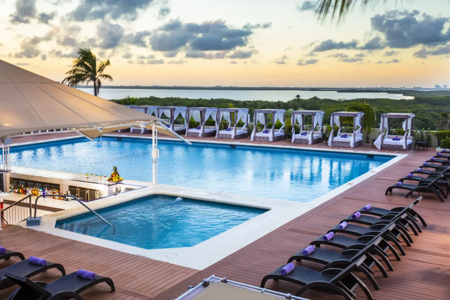 Pool deck at Crown Paradise Club Cancun with cabanas, loungers, bar, and lagoon view at sunset.