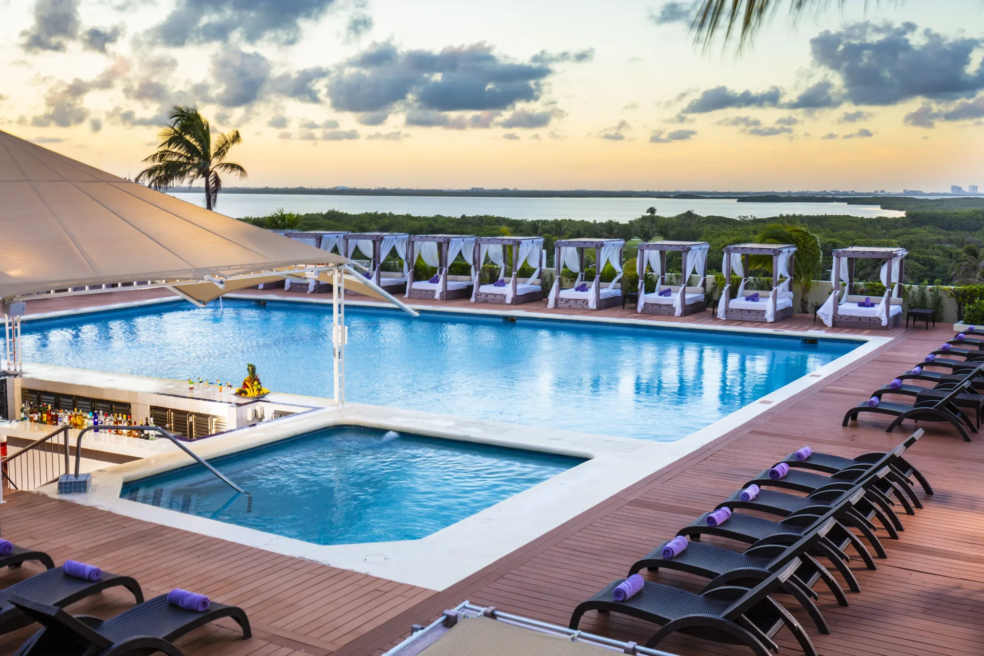 Pool deck at Crown Paradise Club Cancun with cabanas, loungers, bar, and lagoon view at sunset.