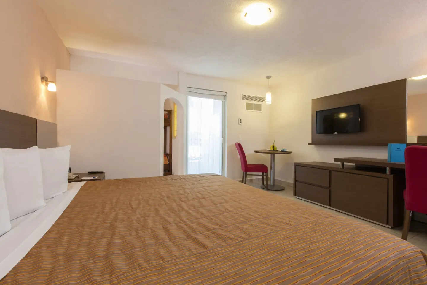 Guest room with king bed, TV wall, red chair and balcony door at Crown Paradise Club Puerto Vallarta.