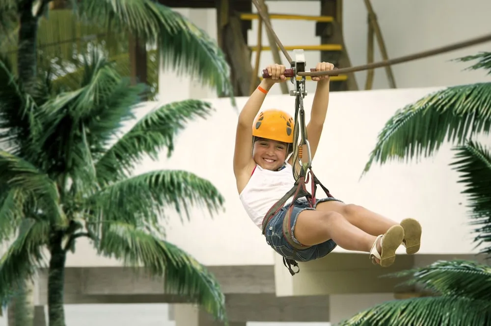 Child riding a zip line between palm trees at Crown Paradise Club Puerto Vallarta.