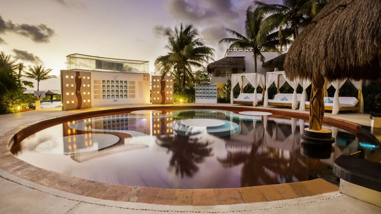 Red-tiled round pool with cabanas and palms at dusk at Desire Pearl Riviera Maya Resort.
