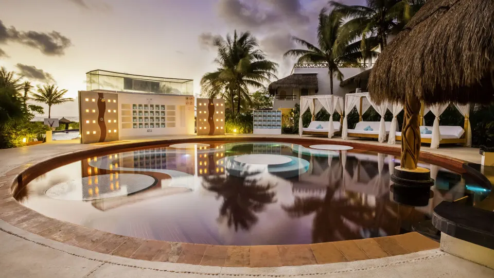 Red-tiled round pool with cabanas and palms at dusk at Desire Pearl Riviera Maya Resort.