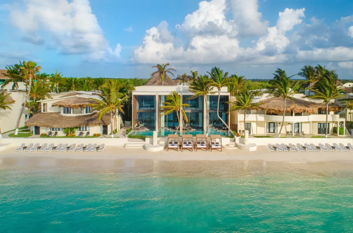 Beachfront buildings, loungers, palms, and turquoise water at Desire Riviera Maya Resort.