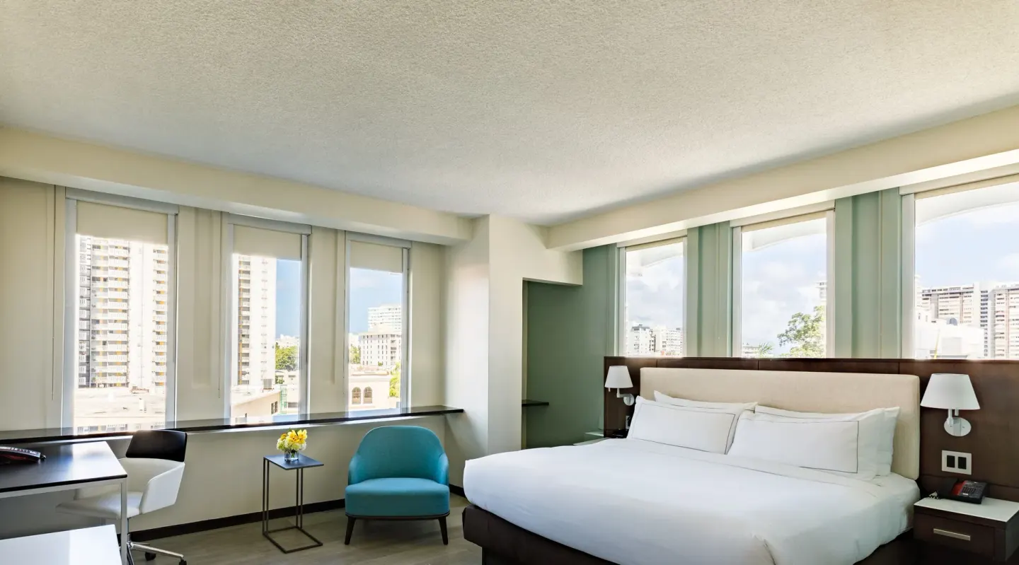 King bed corner room with multiple windows, desk, teal chair, and city views at DoubleTree San Juan.