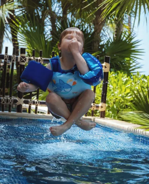 Child jumping into a pool at Dreams Riviera Cancun Resort & Spa with tropical greenery behind.
