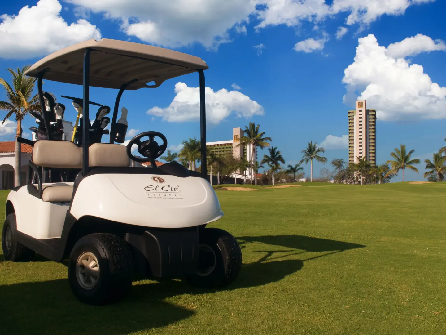 Golf cart on a green course near resort towers at El Cid Castilla Beach Hotel.
