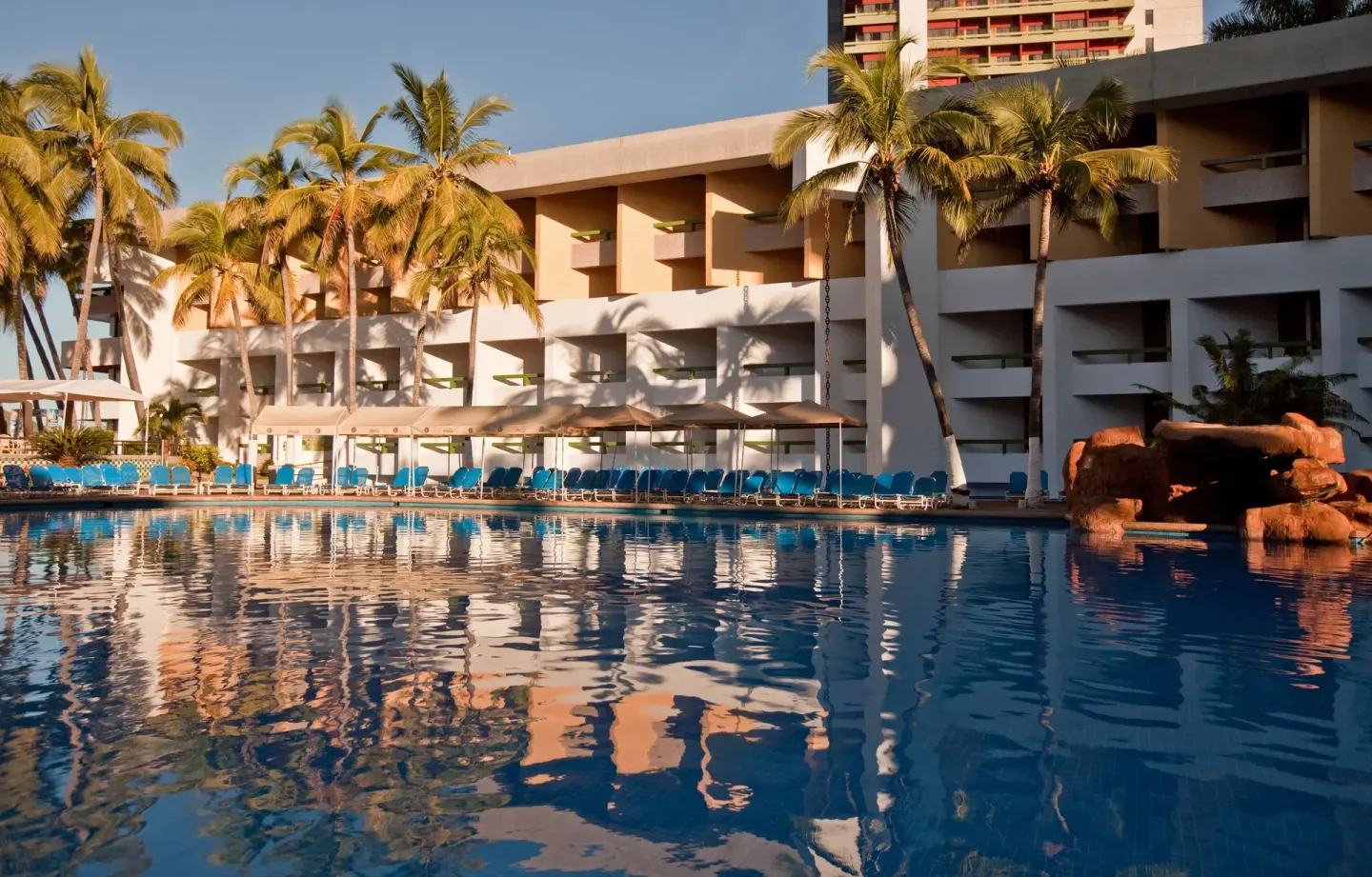 Poolside guest building and palm trees reflected in water at El Cid Castilla Beach Hotel.