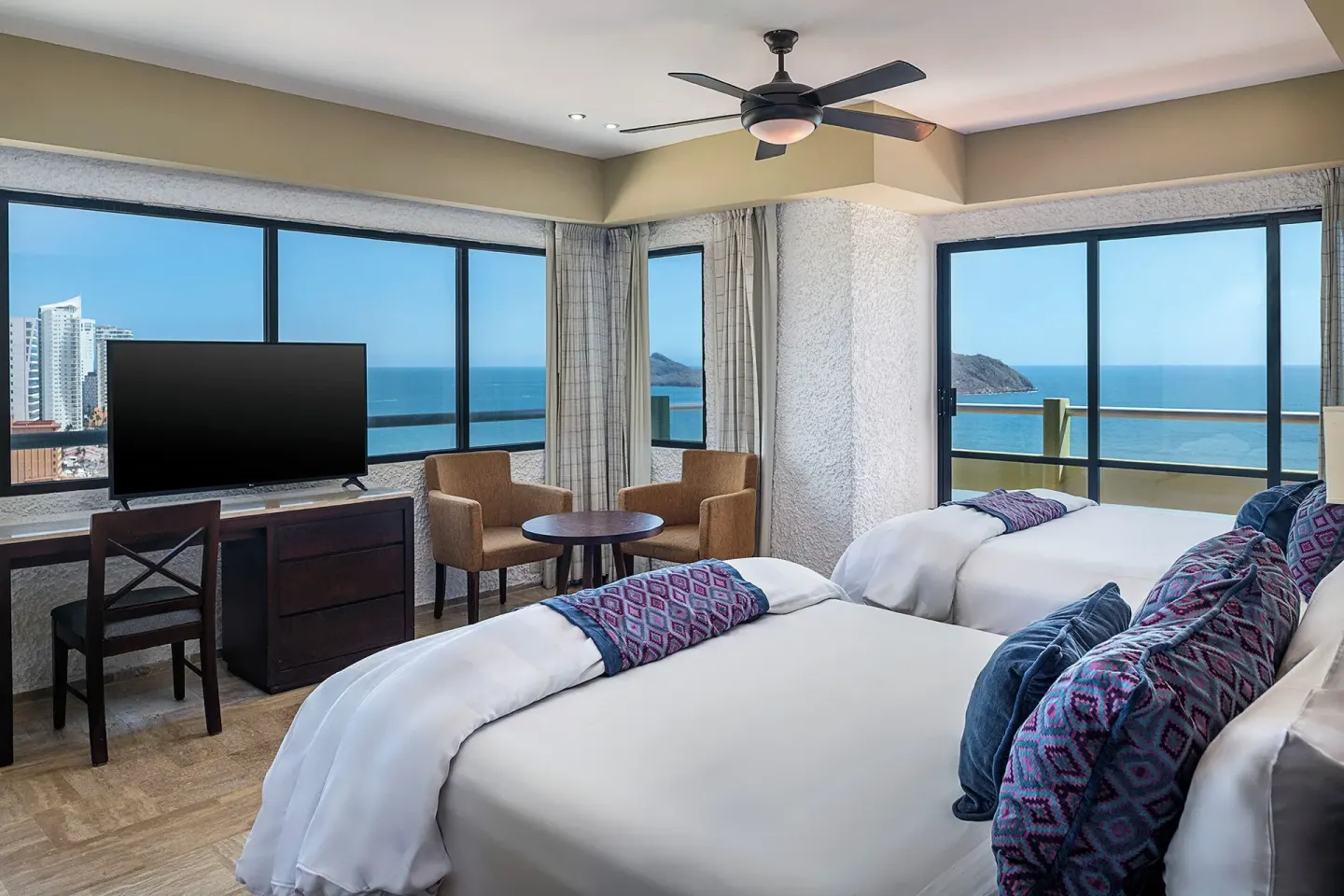 One Bedroom Suite with two beds, TV, seating, and ocean-view windows at El Cid El Moro Beach Hotel.