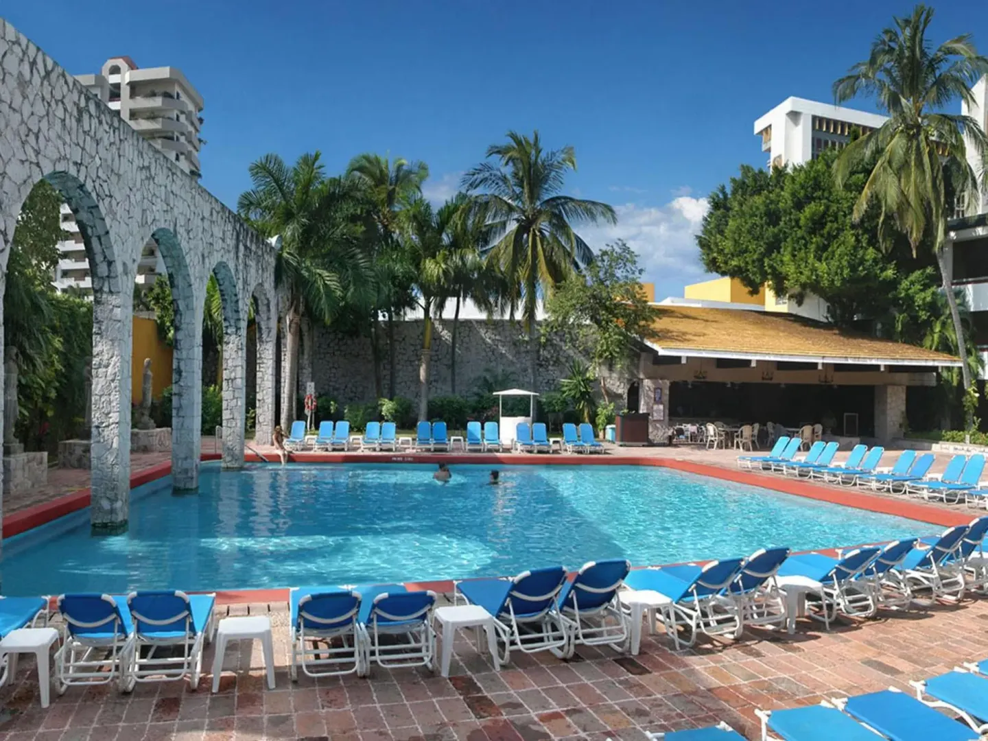 Pool deck with blue loungers, stone arches and palms at El Cid Granada Hotel.