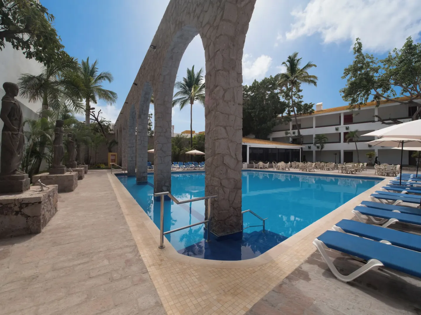 Pool deck with stone arches, statues, and blue loungers at El Cid Granada Hotel.