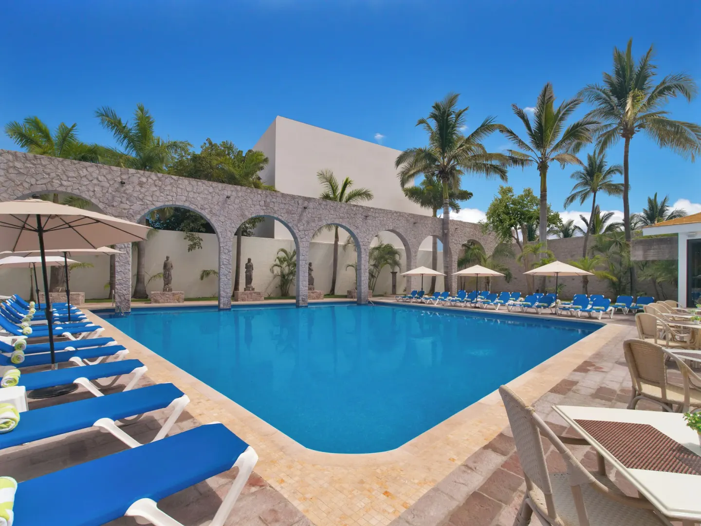 Pool with blue loungers, umbrellas, stone arches and palms at El Cid Granada Hotel.