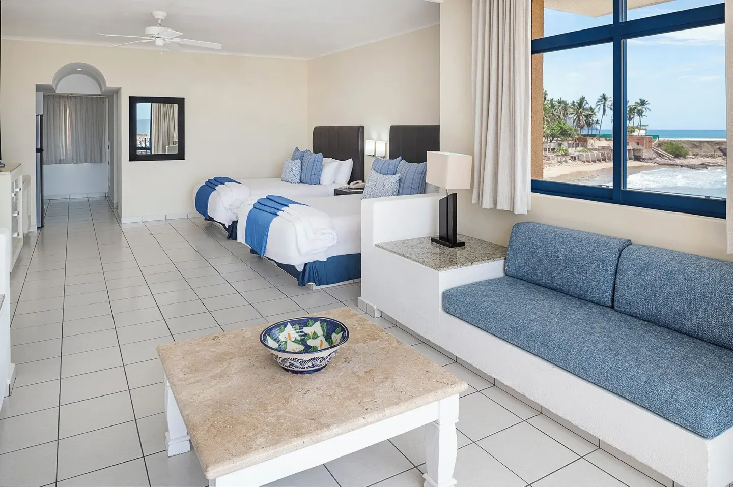 Junior Suite with two beds, blue sofa, and ocean-facing window at El Cid Marina Beach Hotel.
