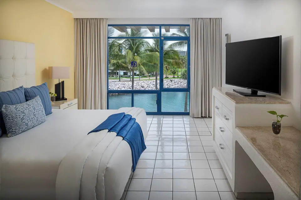 One Bedroom Suite king bed facing blue-framed windows at El Cid Marina Beach Hotel.