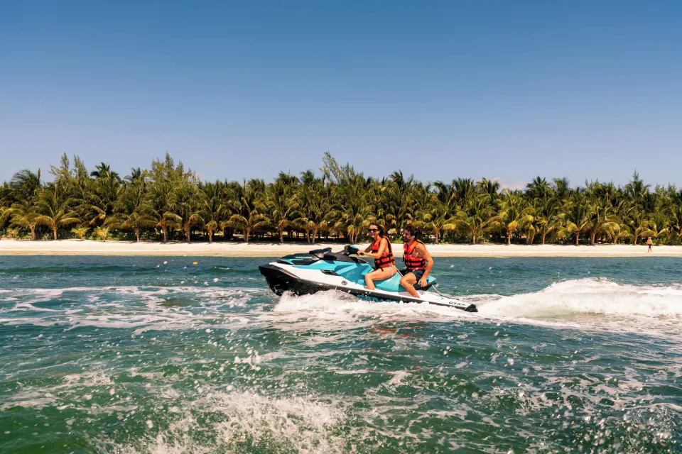 Two people riding a jet ski near a palm-lined beach at El Dorado Seaside Suites