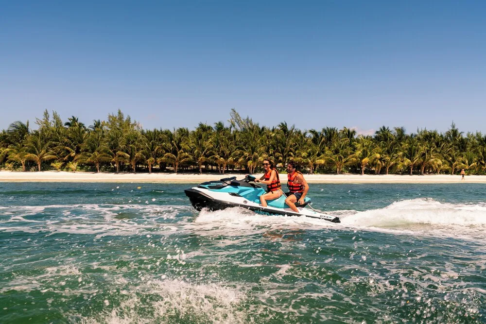 Two people riding a jet ski near the palm-lined shore of El Dorado Seaside Suites