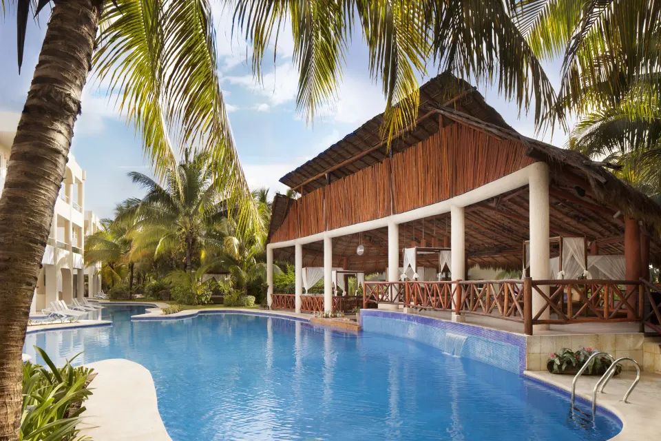 Resort pool with palapa-roofed dining area and palm trees at El Dorado Seaside Suites