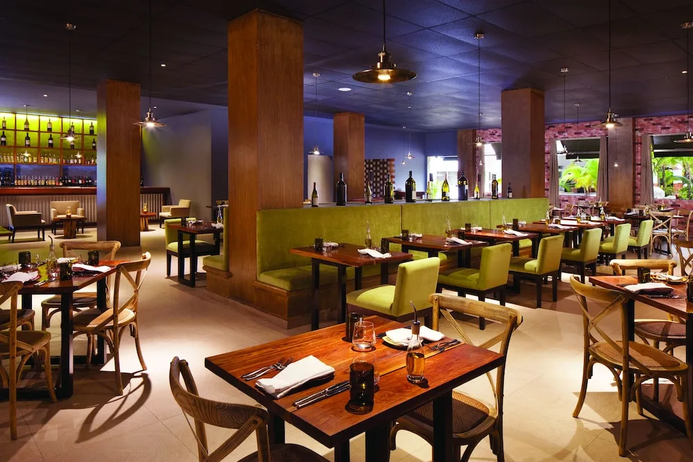 Interior of a resort restaurant with wooden tables and green banquette seating at El Dorado Seaside Suites