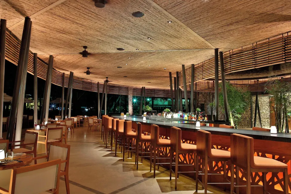 Long illuminated bar with high stools under a woven ceiling at El Mangroove Papagayo