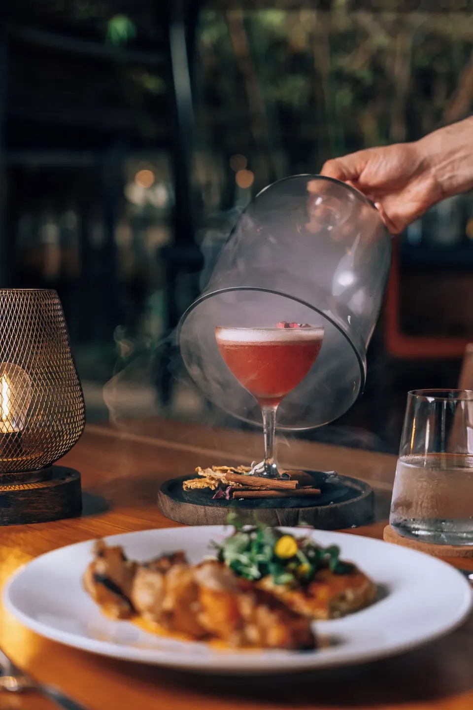 Smoked cocktail being revealed next to plated food at El Mangroove Papagayo