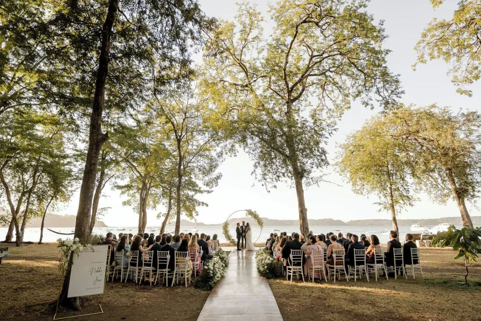 Wedding ceremony with guests seated under trees facing the ocean at El Mangroove Papagayo