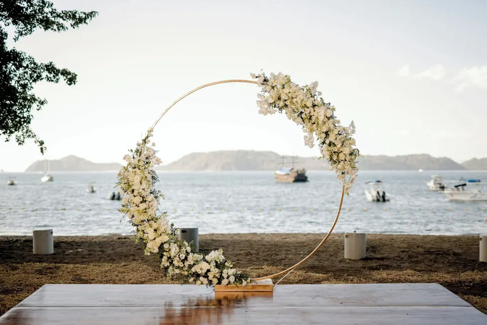 Circular floral wedding arch on the sand with boats and ocean in the background at El Mangroove Papagayo