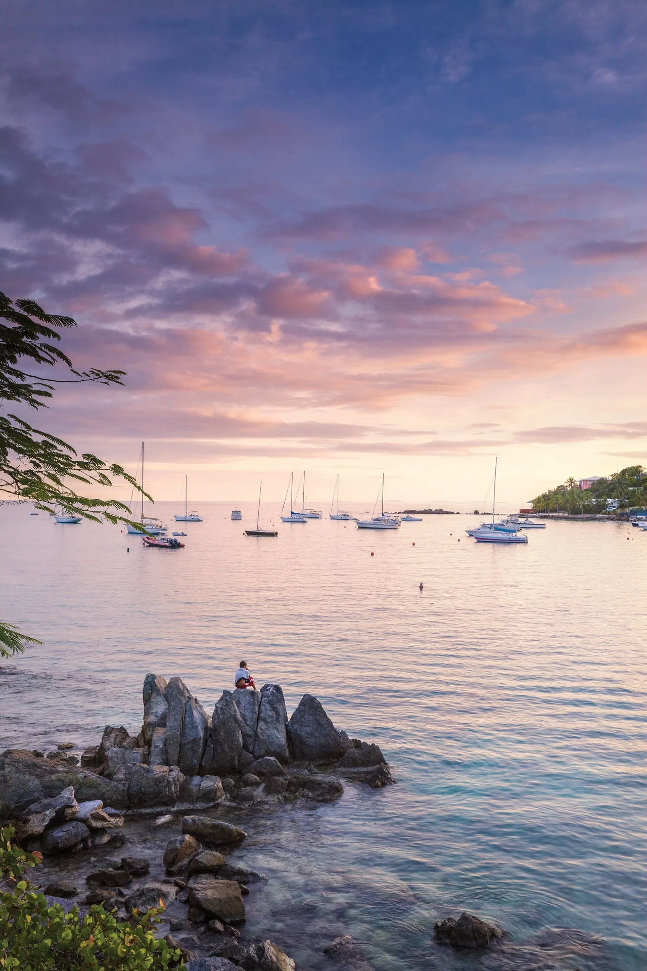 Pastel sunset over a rocky shoreline and sailboats at Elysian Beach Resort.