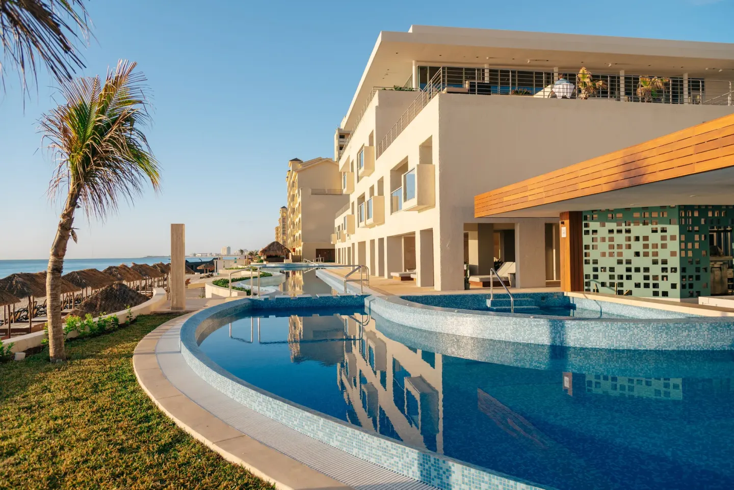 Oceanfront pool area with curved basins, resort building, palm tree, and beach beyond at Emporio Cancun.