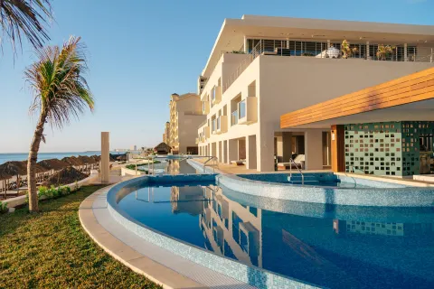 Oceanfront pool area with curved basins, resort building, palm tree, and beach beyond at Emporio Cancun.