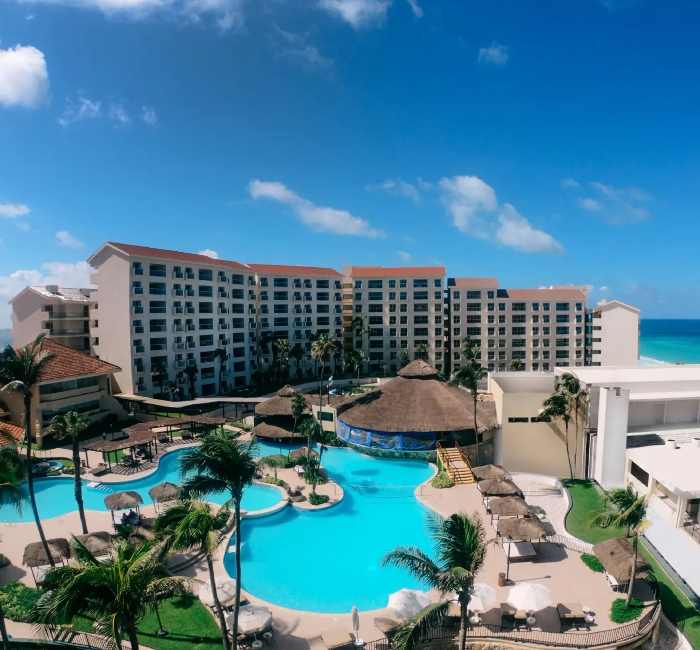 Pool courtyard, palms, resort buildings, and ocean are seen from above at Emporio Cancun.