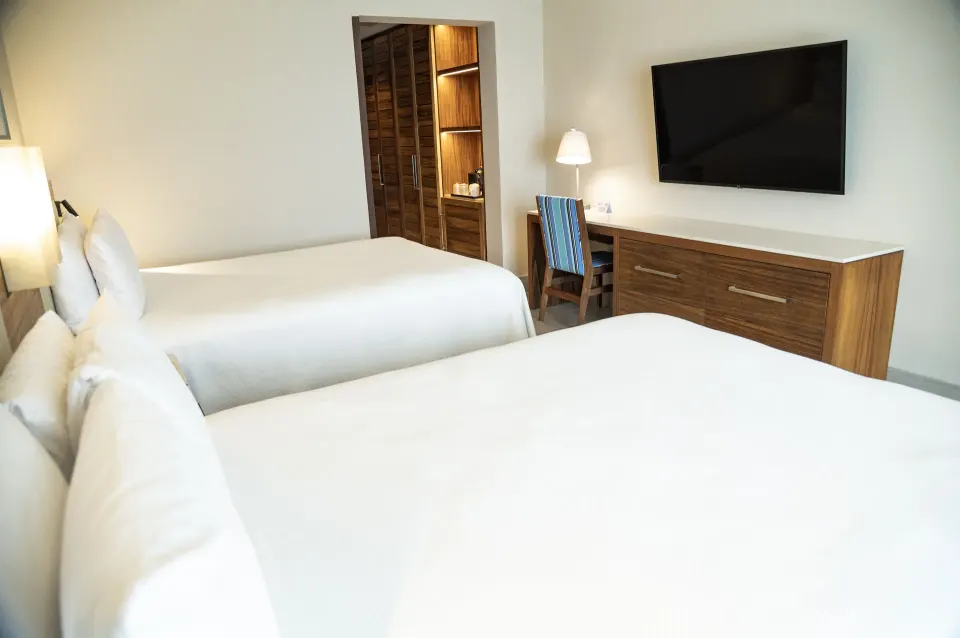 Two beds in a Swim Up Oceanfront room at Emporio Cancun facing a wall-mounted TV and desk.