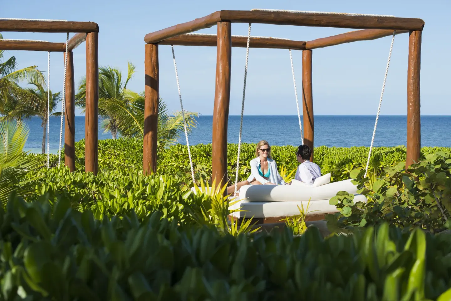 Couple seated on a hanging daybed amid greenery by the ocean at Excellence Playa Mujeres.