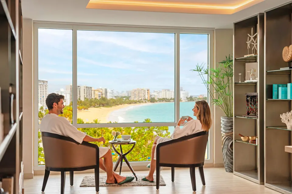 Guests relax in chairs by a large beach-view window at Fairmont El San Juan.