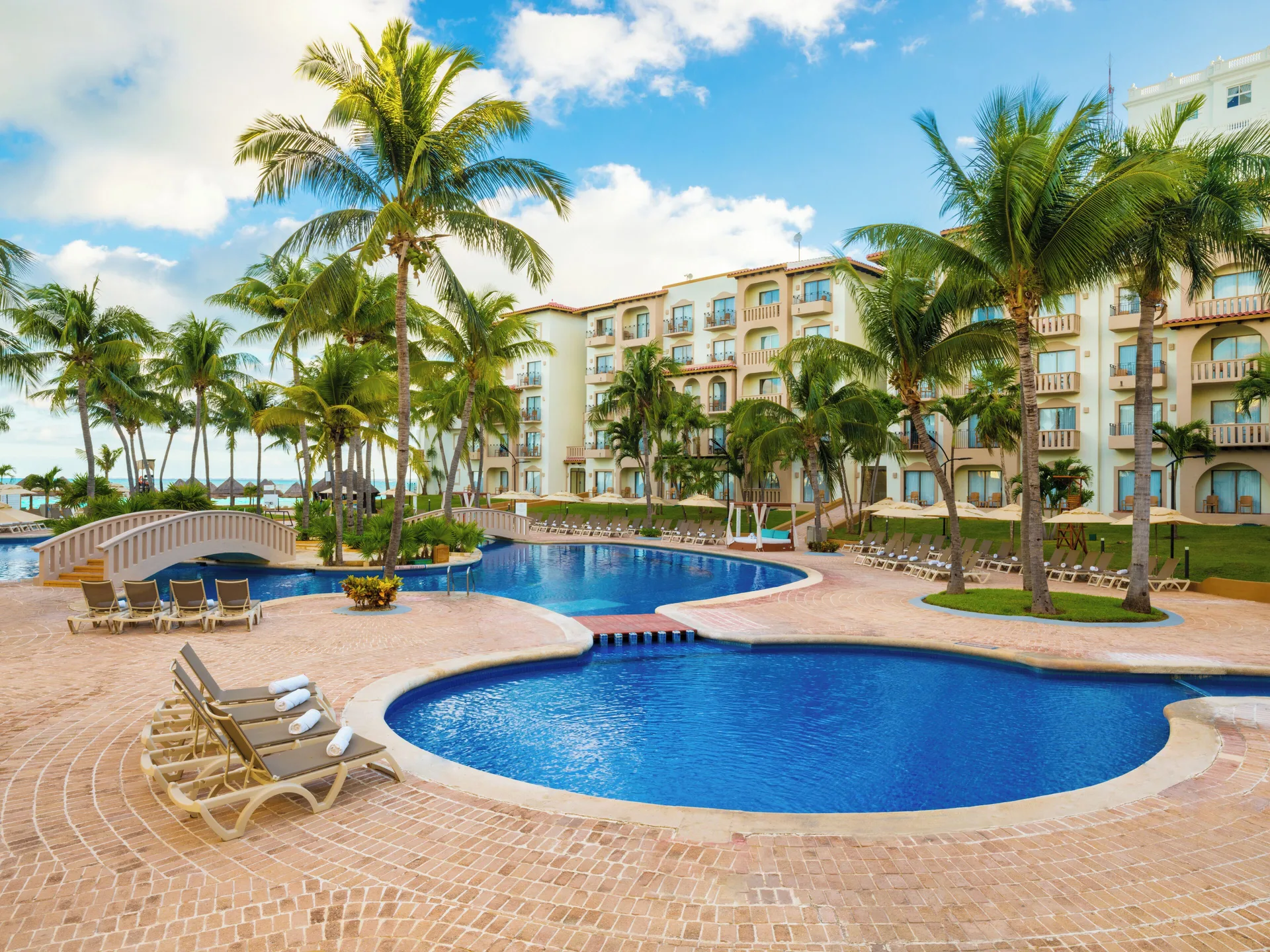 Curved pools, bridge, loungers, and resort buildings at Fiesta Americana Cancun Villas.