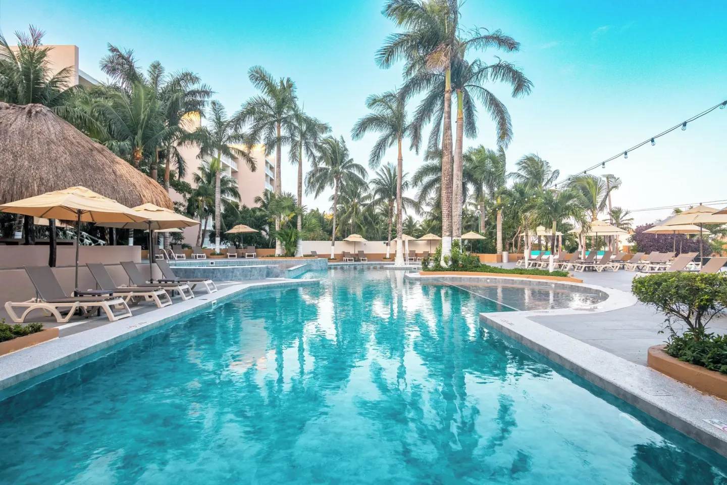 Long resort pool with loungers, palms, and a thatched-roof area at Fiesta Americana Cozumel.
