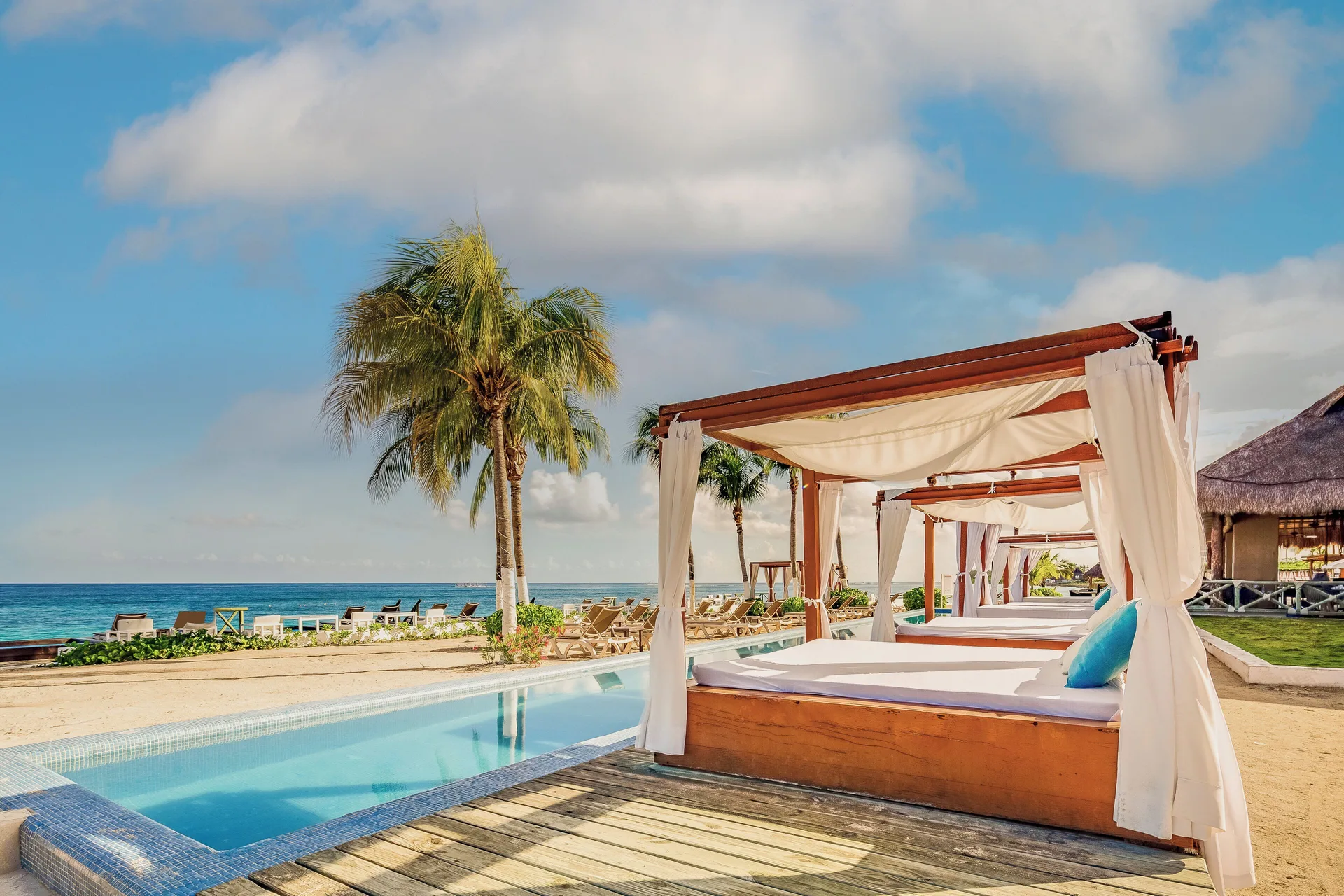 Poolside daybeds with white curtains sit beside the beach at Fiesta Americana Cozumel.