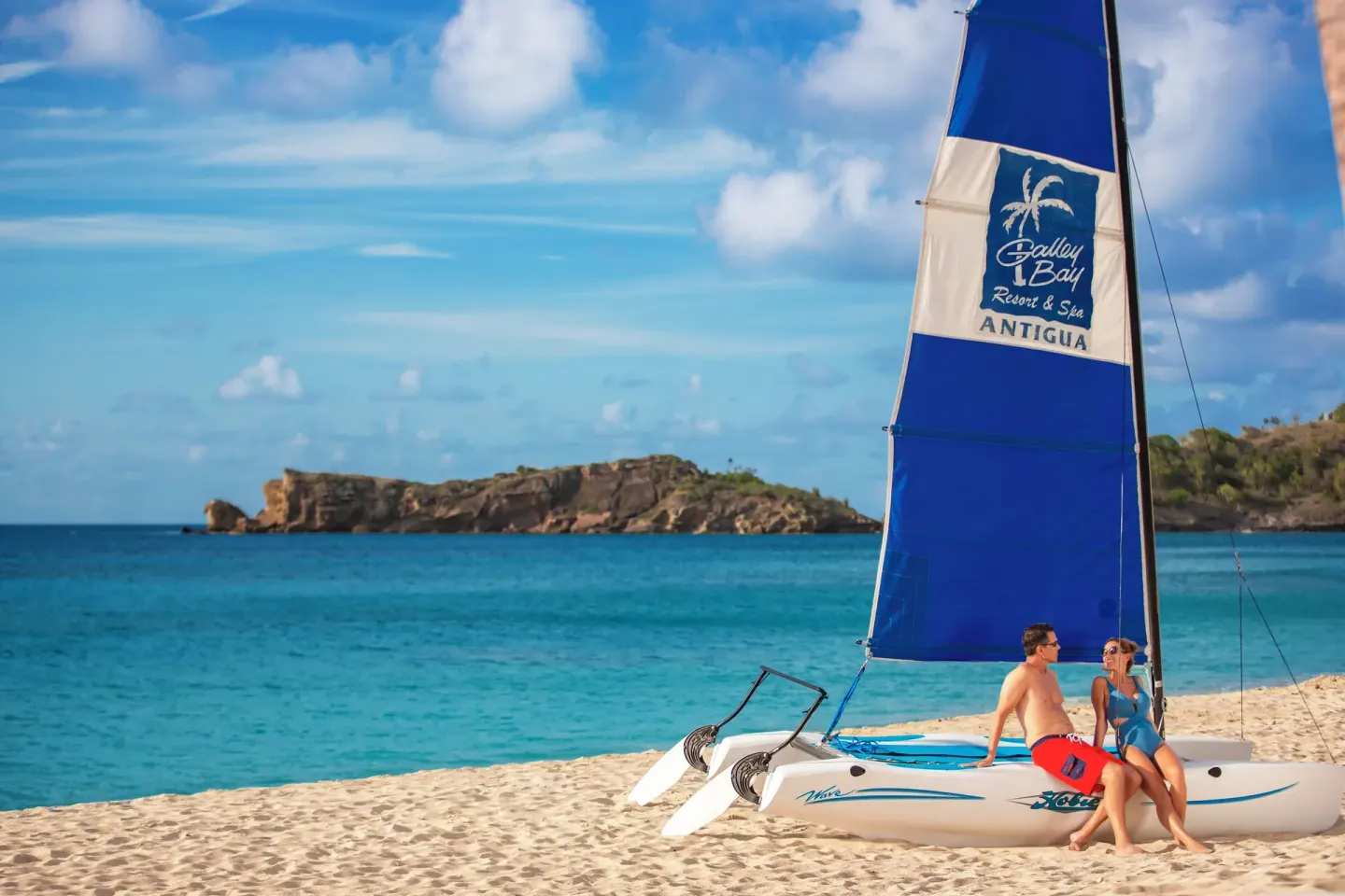 A couple sits on a beached catamaran with a branded sail at Galley Bay Resort & Spa.