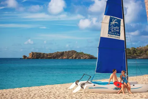 A couple sits on a beached catamaran with a branded sail at Galley Bay Resort & Spa.
