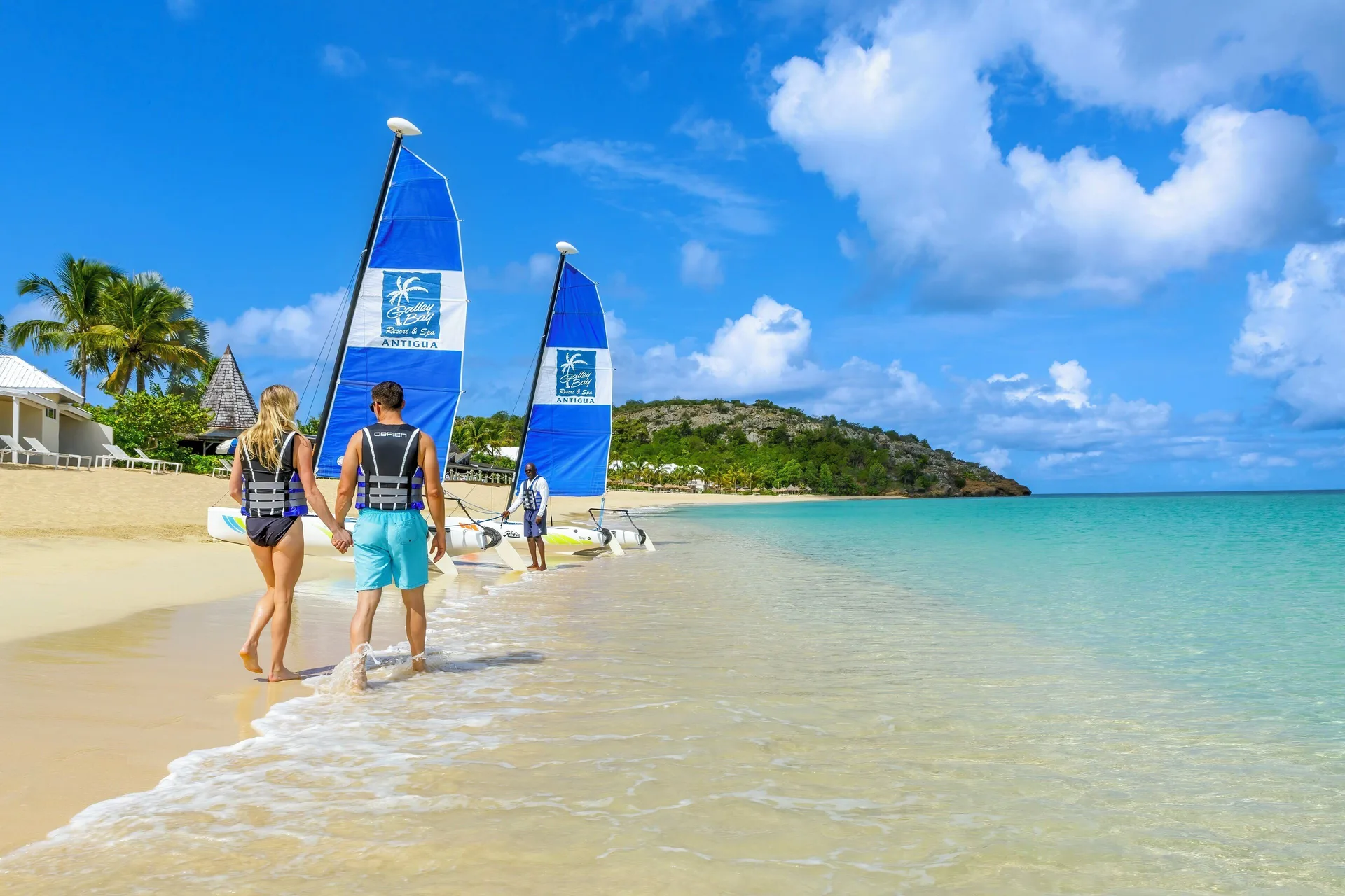 A couple walks along the beach toward blue-sailed catamarans at Galley Bay Resort & Spa.