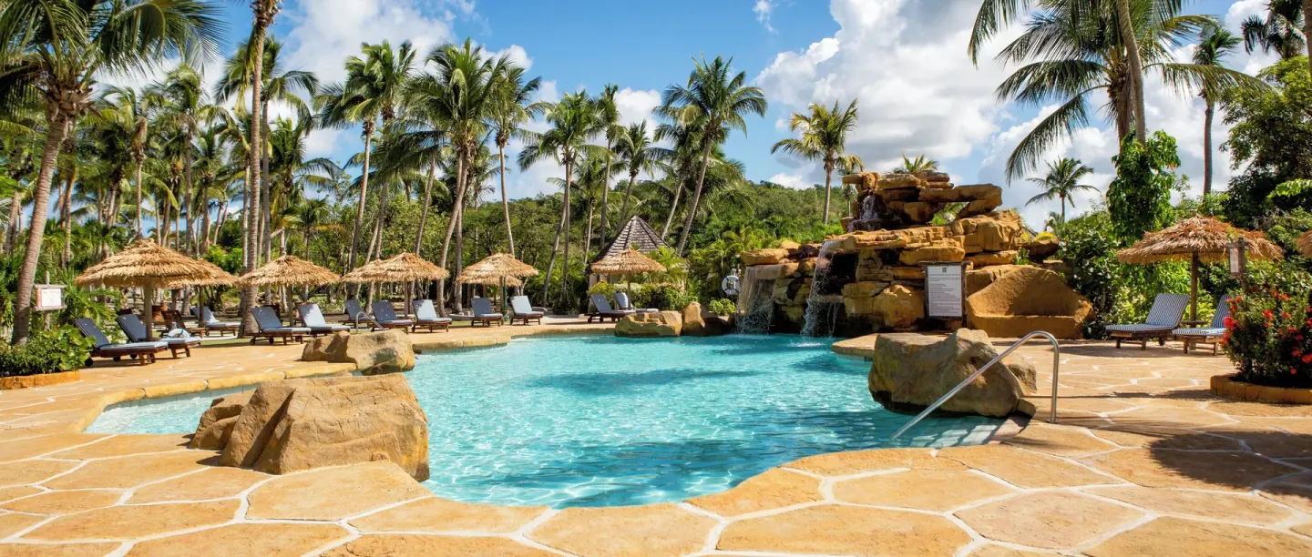 Curved resort pool with rock waterfall, loungers, thatched umbrellas, and palms at Galley Bay Resort & Spa.