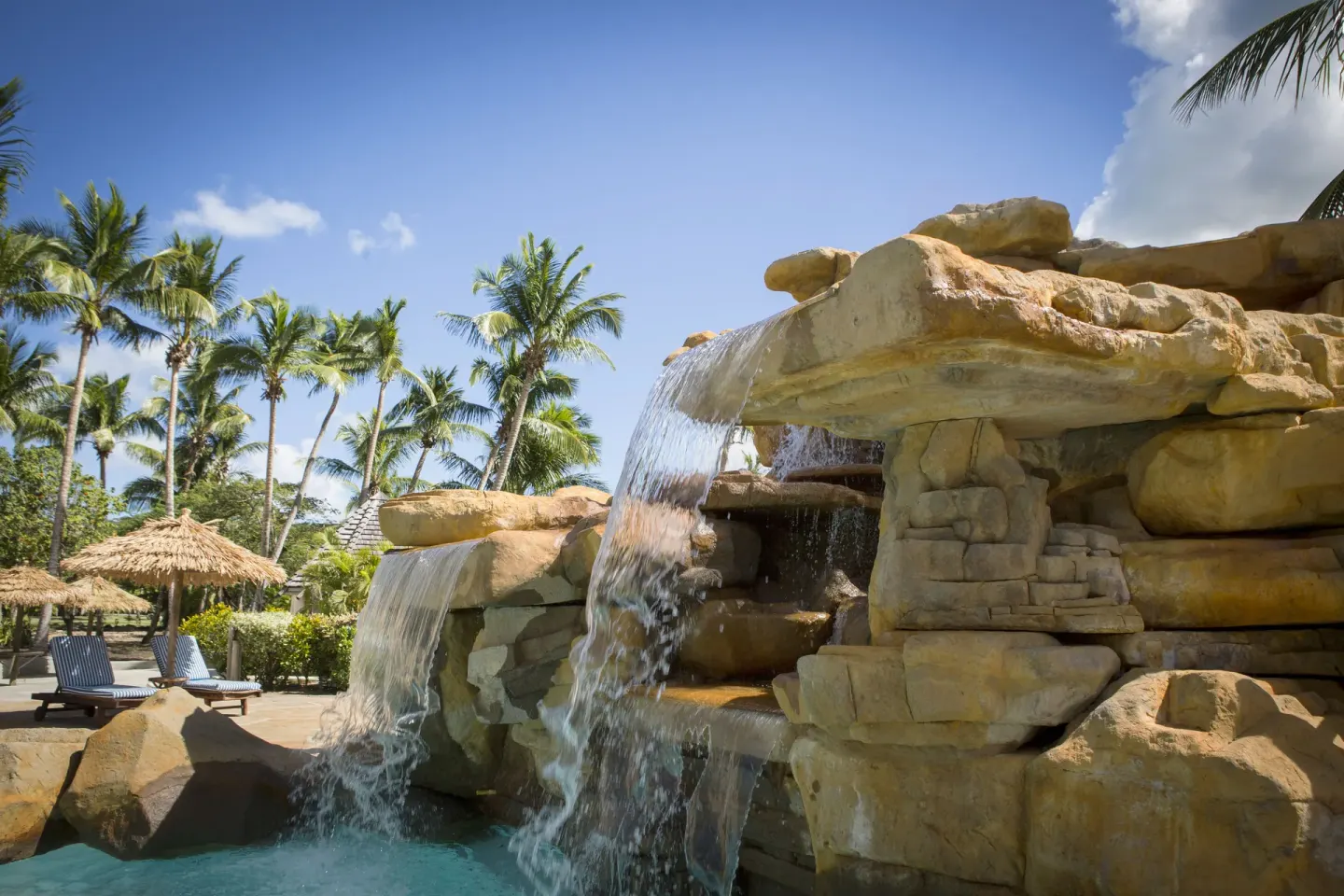 Rock waterfall pours into the pool at Galley Bay Resort & Spa, with palms and loungers nearby.