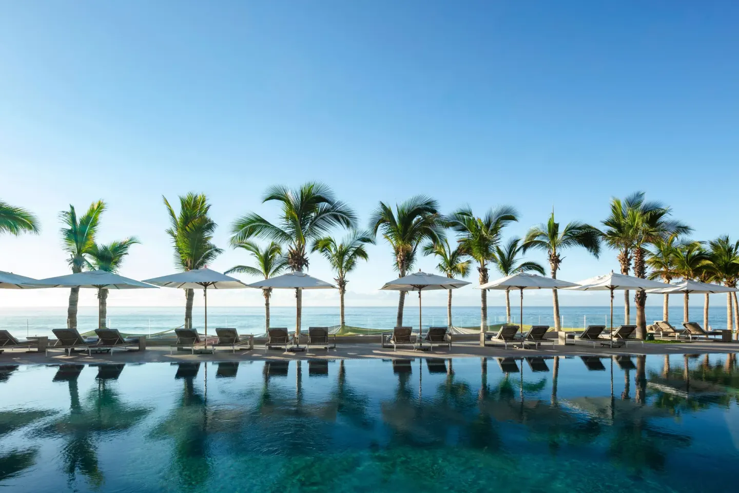 Pool lined with palms, umbrellas, loungers, and ocean beyond at Garza Blanca Los Cabos.