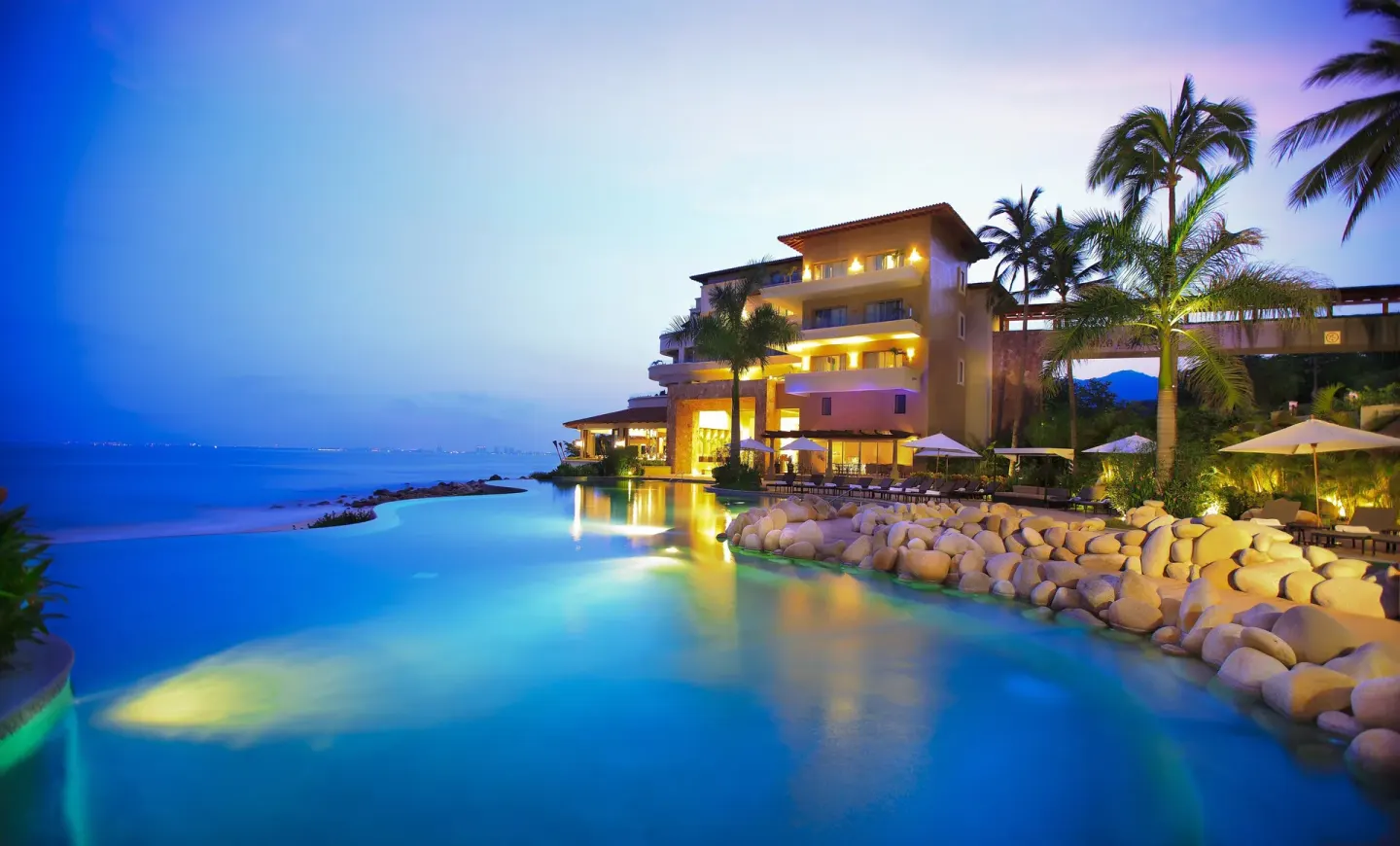 Illuminated beachfront pool at dusk at Garza Blanca Preserve Resort & Spa with palm trees and loungers.
