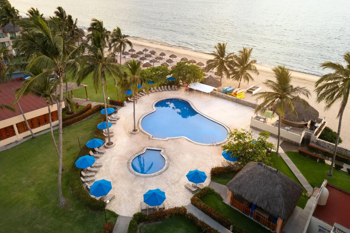 Aerial beachside pool with palms, blue umbrellas, and sand beyond at Grand Decameron Complex Bucerias.