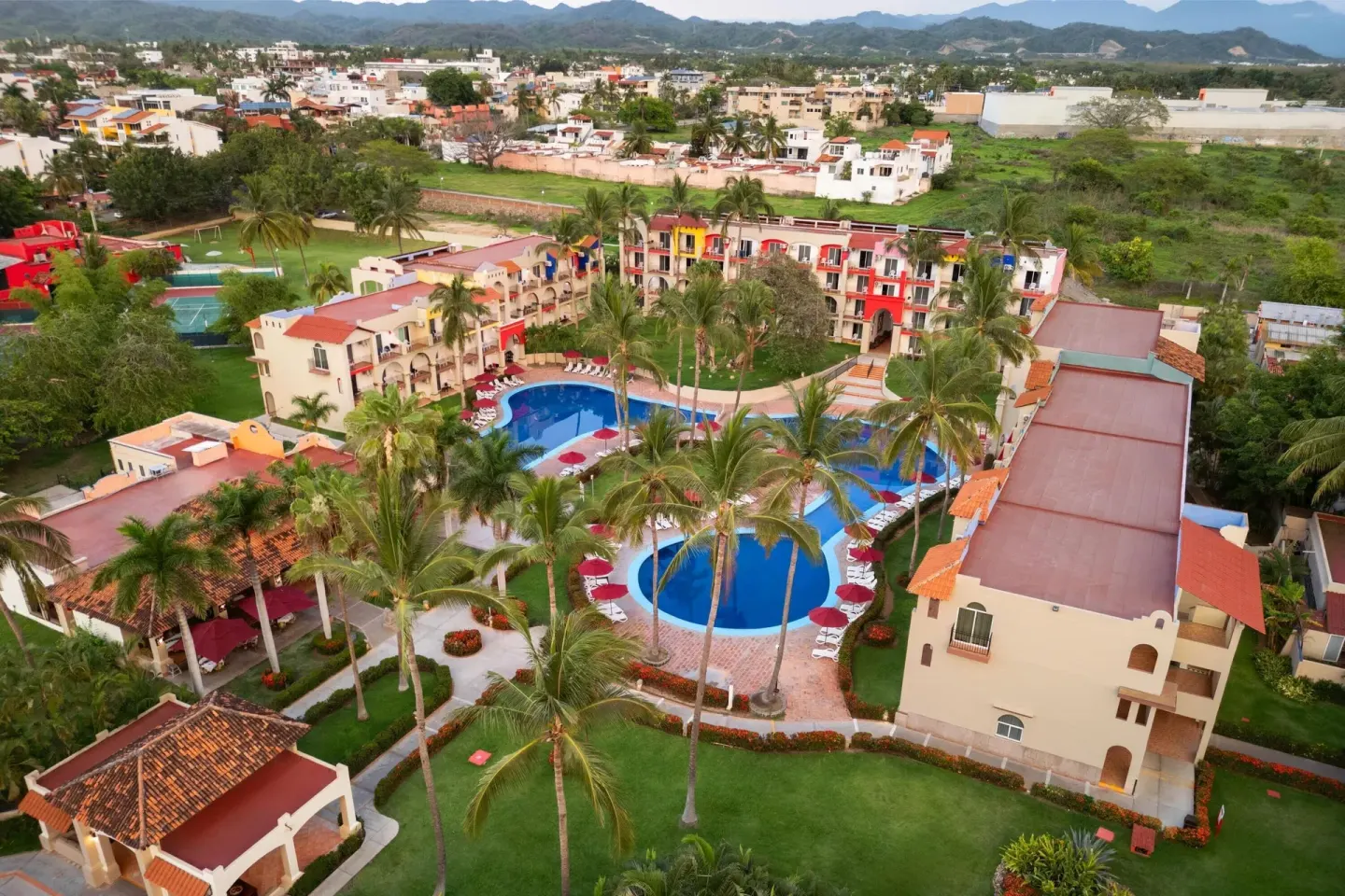 Aerial resort grounds with pools, palms, colorful buildings, and mountains at Grand Decameron Complex Bucerias.