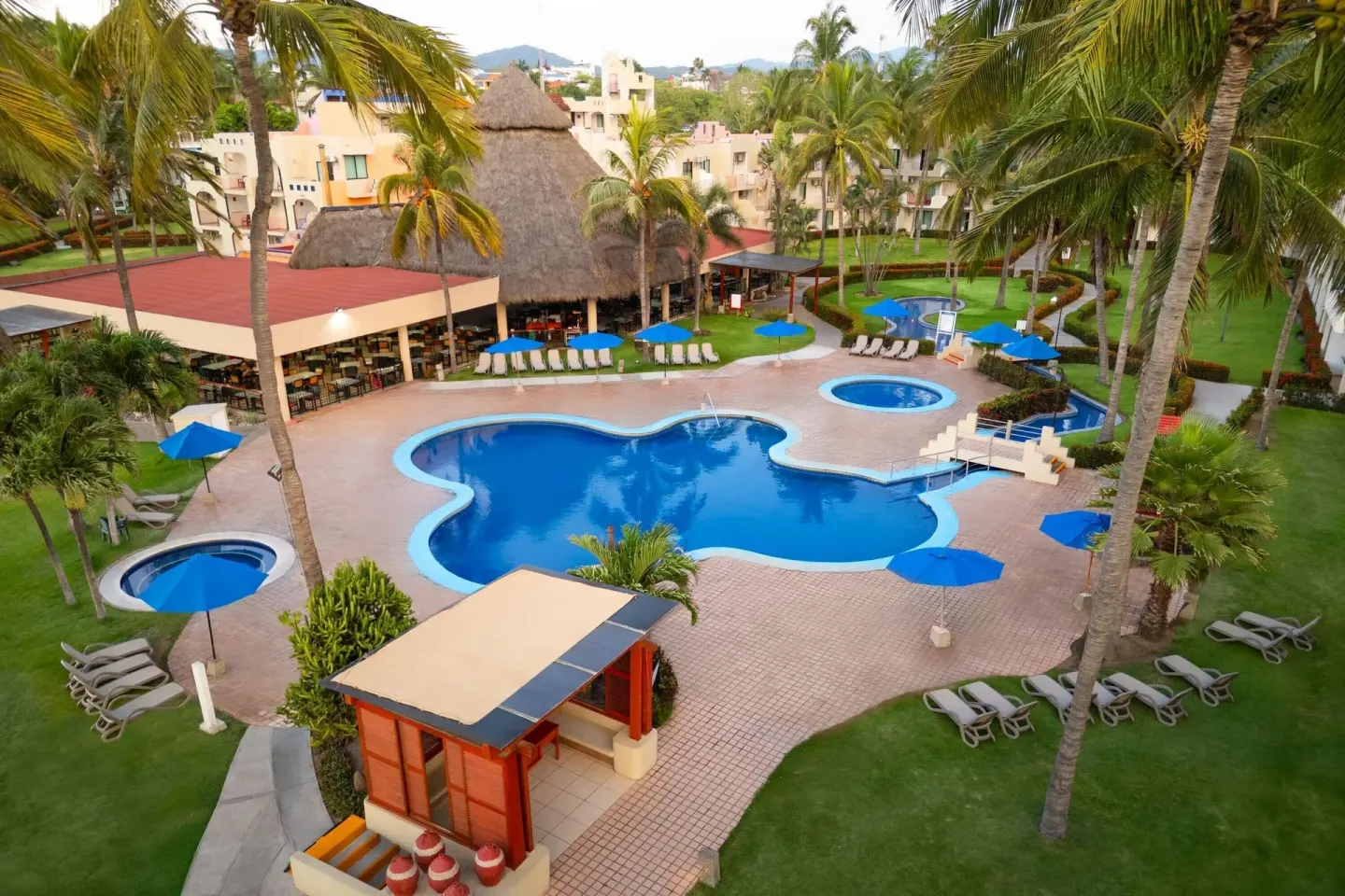Pool courtyard with loungers, blue umbrellas, palms, and thatched roofs at Grand Decameron Complex Bucerias.