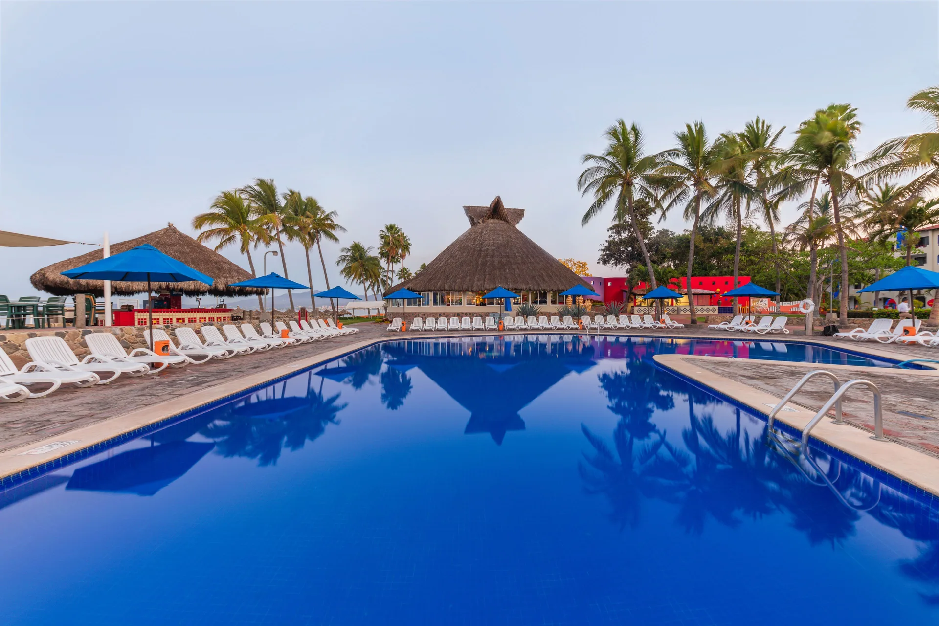 Pool with blue umbrellas, loungers, palms, and palapa roofs at Grand Decameron Complex Bucerias.