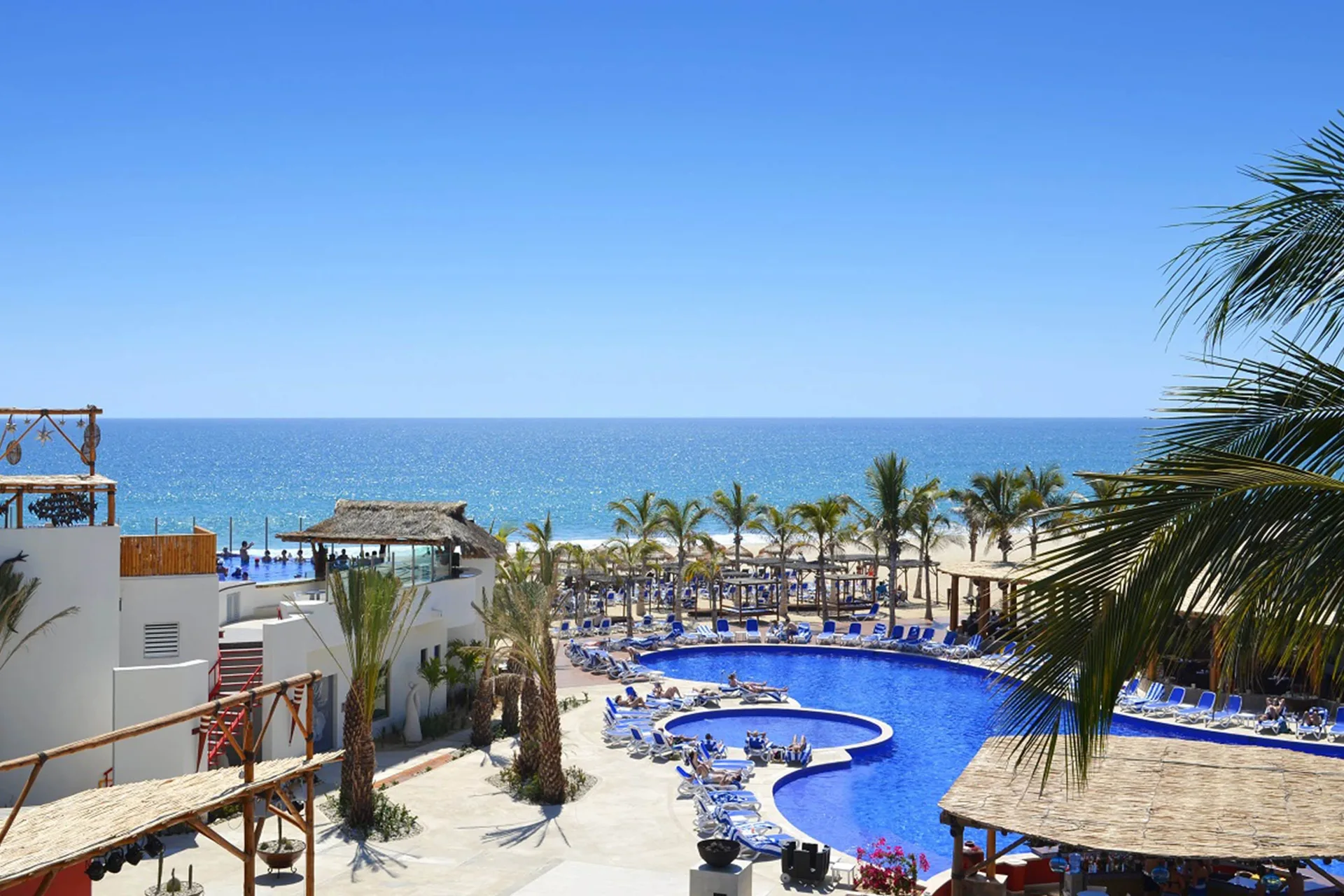 Oceanfront pool with blue loungers and palm trees at Grand Decameron Los Cabos.