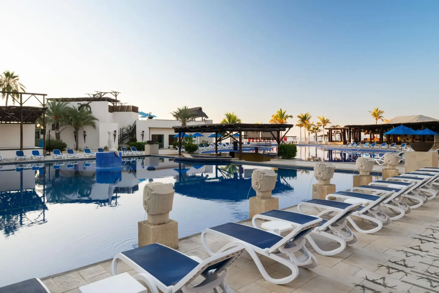 Pool deck with blue loungers, calm water, palms, and ocean beyond at Grand Decameron Los Cabos.