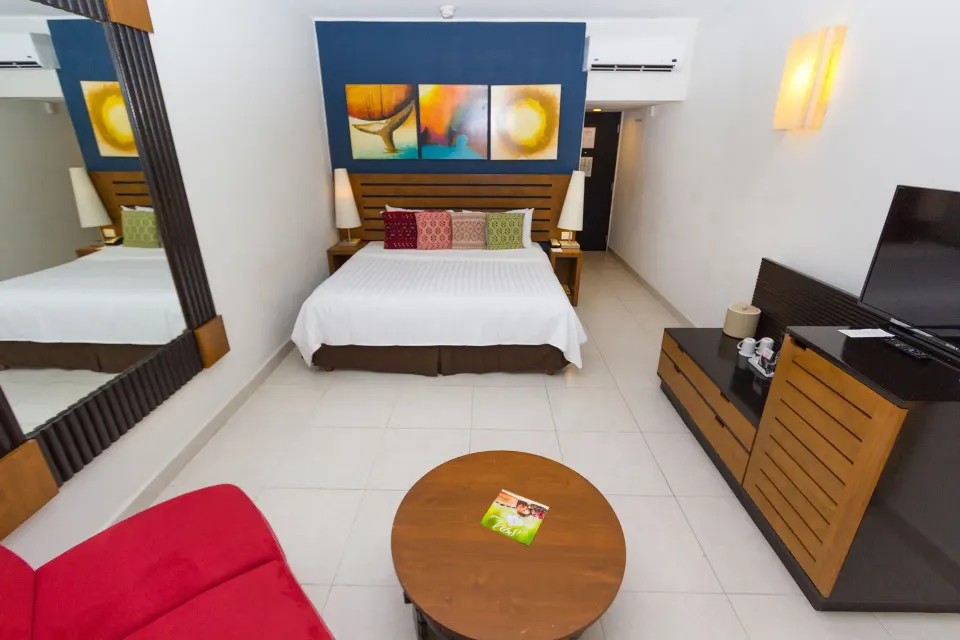 Standard Room with king bed, TV console, red sofa, and large mirror at Grand Decameron Los Cabos.