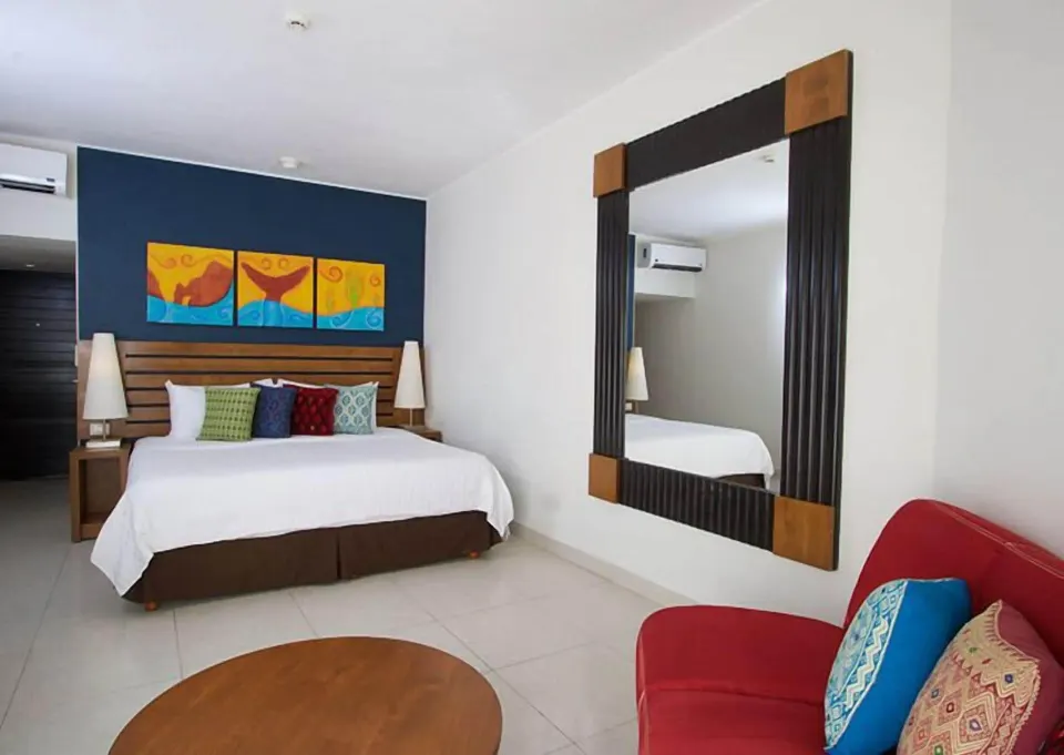 Standard Room with king bed, red sofa, round table, and wall mirror at Grand Decameron Los Cabos.